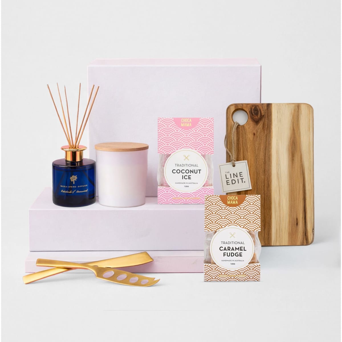 A wooden cheese board, two jars of fudge and coconut ice, a candle, and a diffuser on pink boxes.