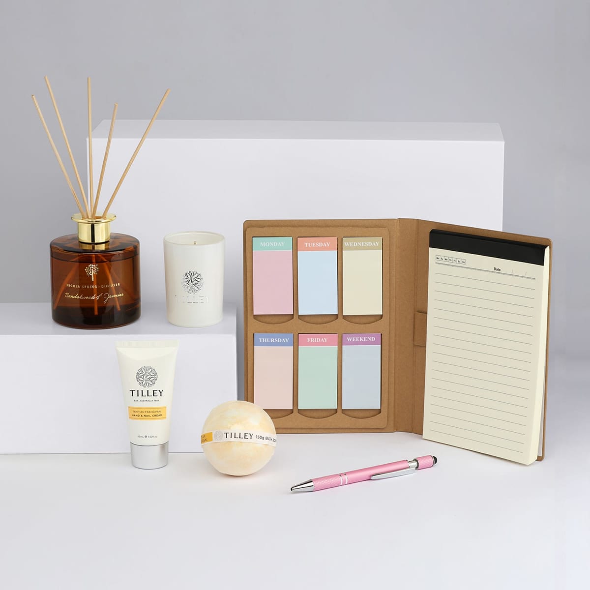 A candle, diffuser, notepad, sticky notes, hand cream, and a pen in soft pastel colours, displayed on a neutral background.