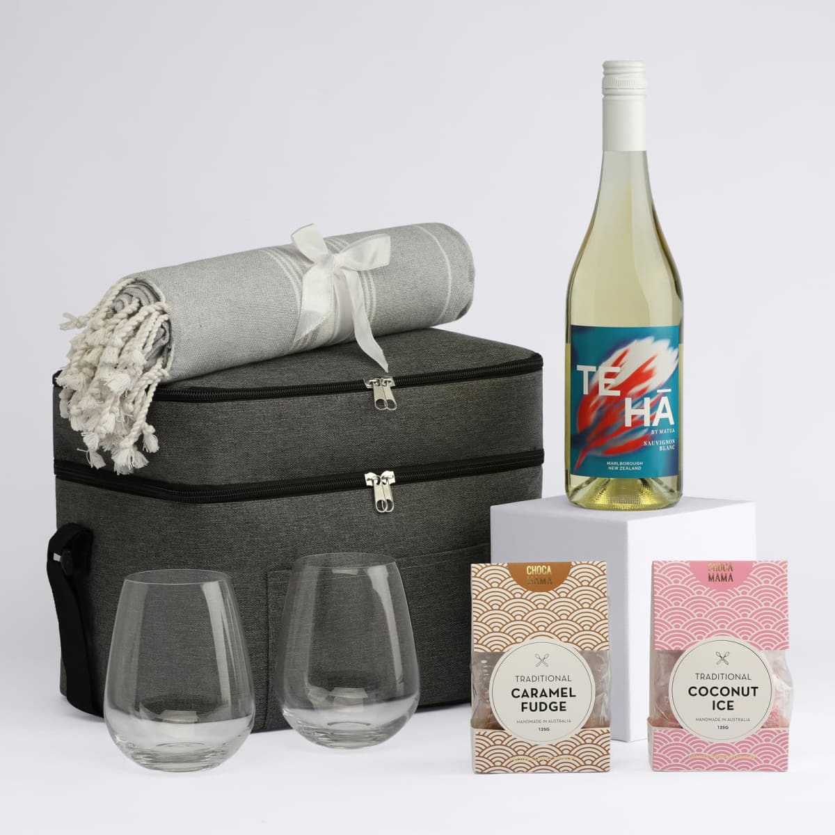A drink bottle and two stemless glasses sit beside a picnic basket, blanket, and two gourmet treats in pink packaging.