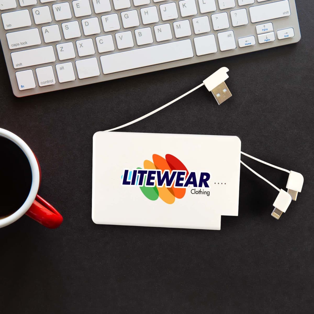 A white power bank with multiple charging cables next to a computer keyboard and a red mug. It has a logo.