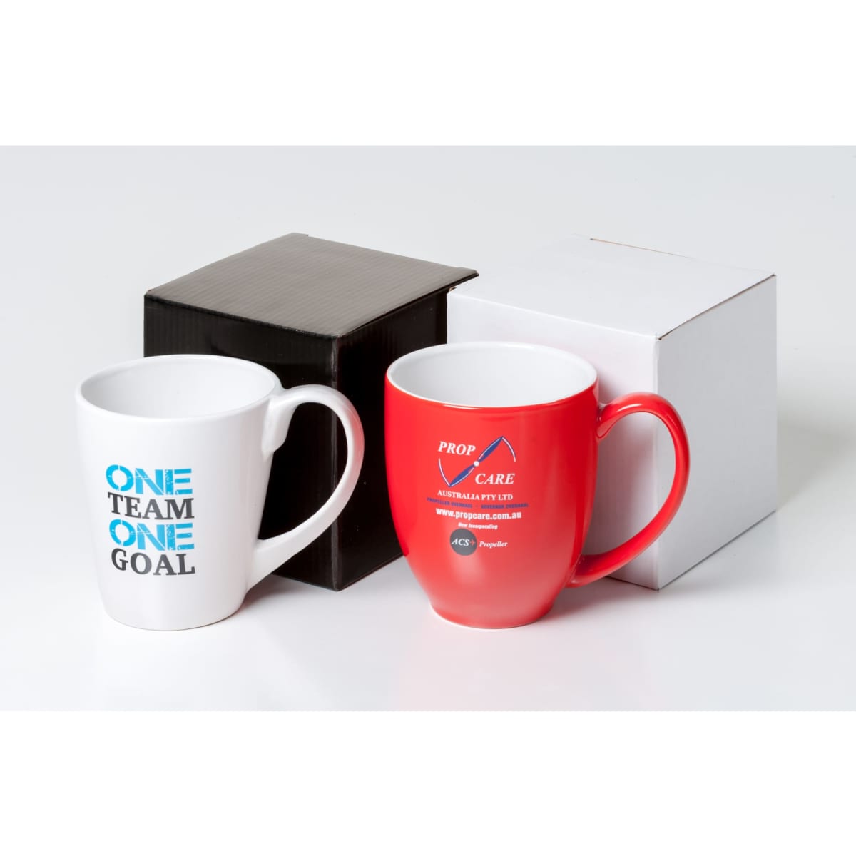 A white mug with blue text and a red mug with a logo, placed beside black and white gift boxes.