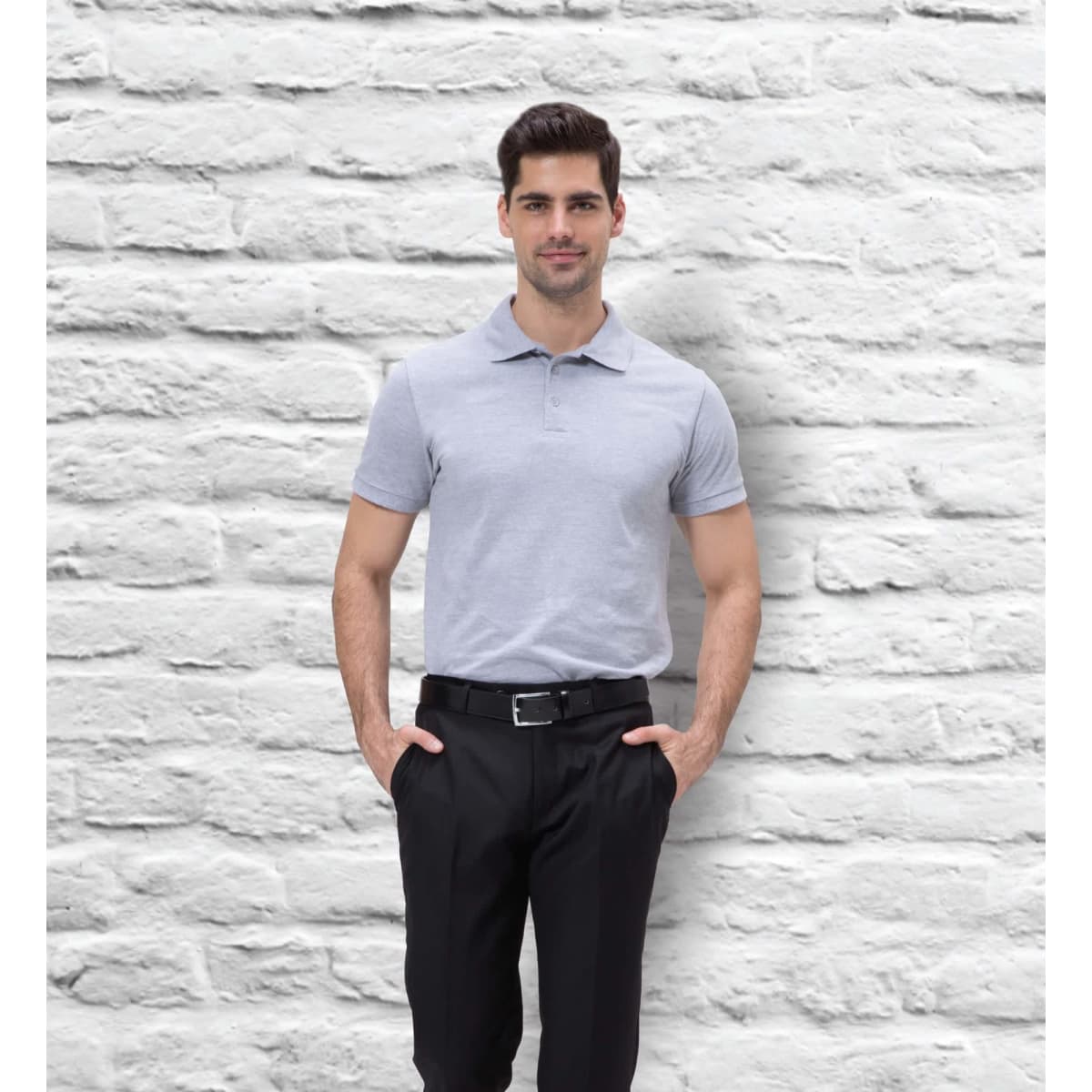 A man in a grey classic polo shirt stands against a textured white brick wall, posing with hands in pockets.