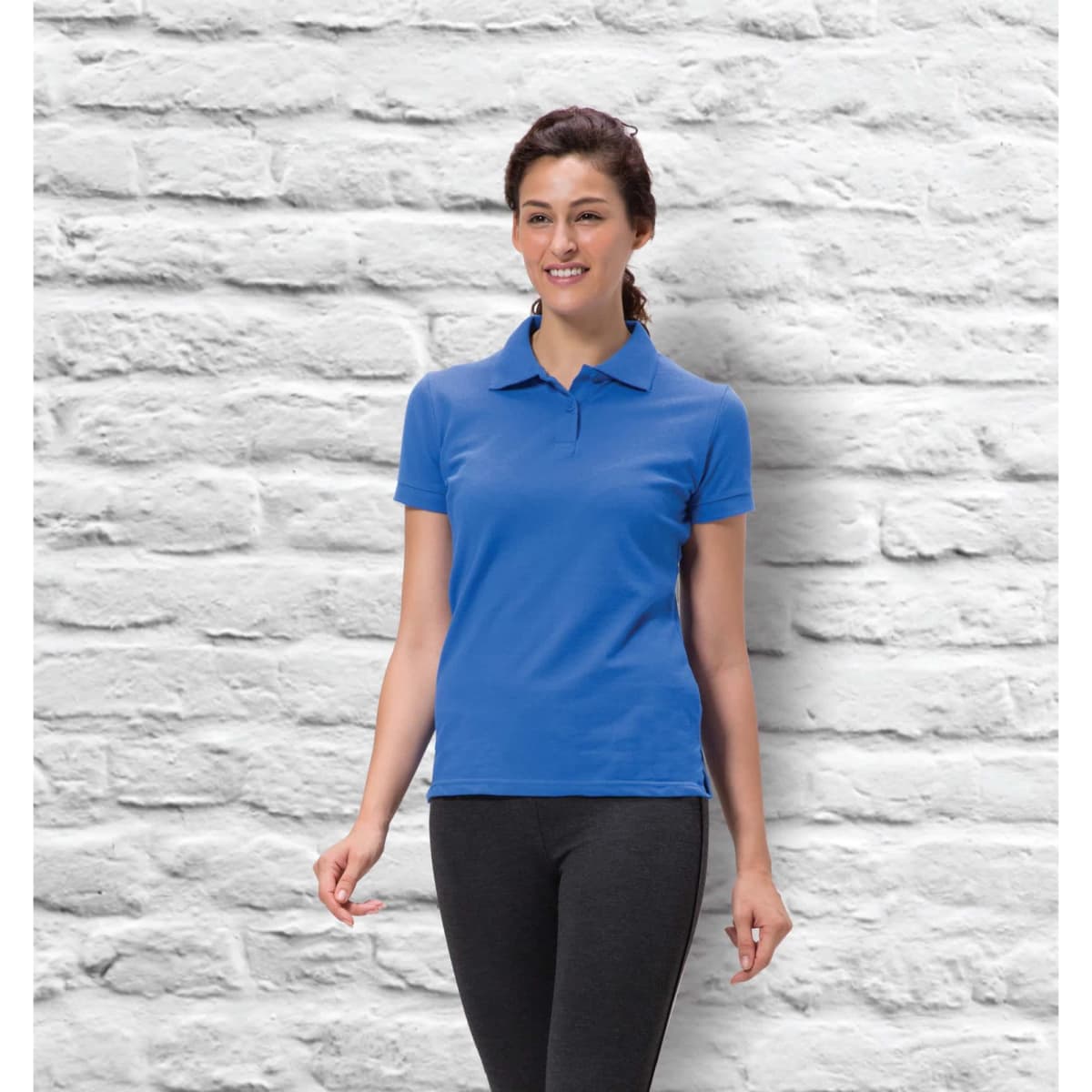A woman in a blue ladies' polo shirt stands against a textured white wall, smiling. The shirt features a logo.