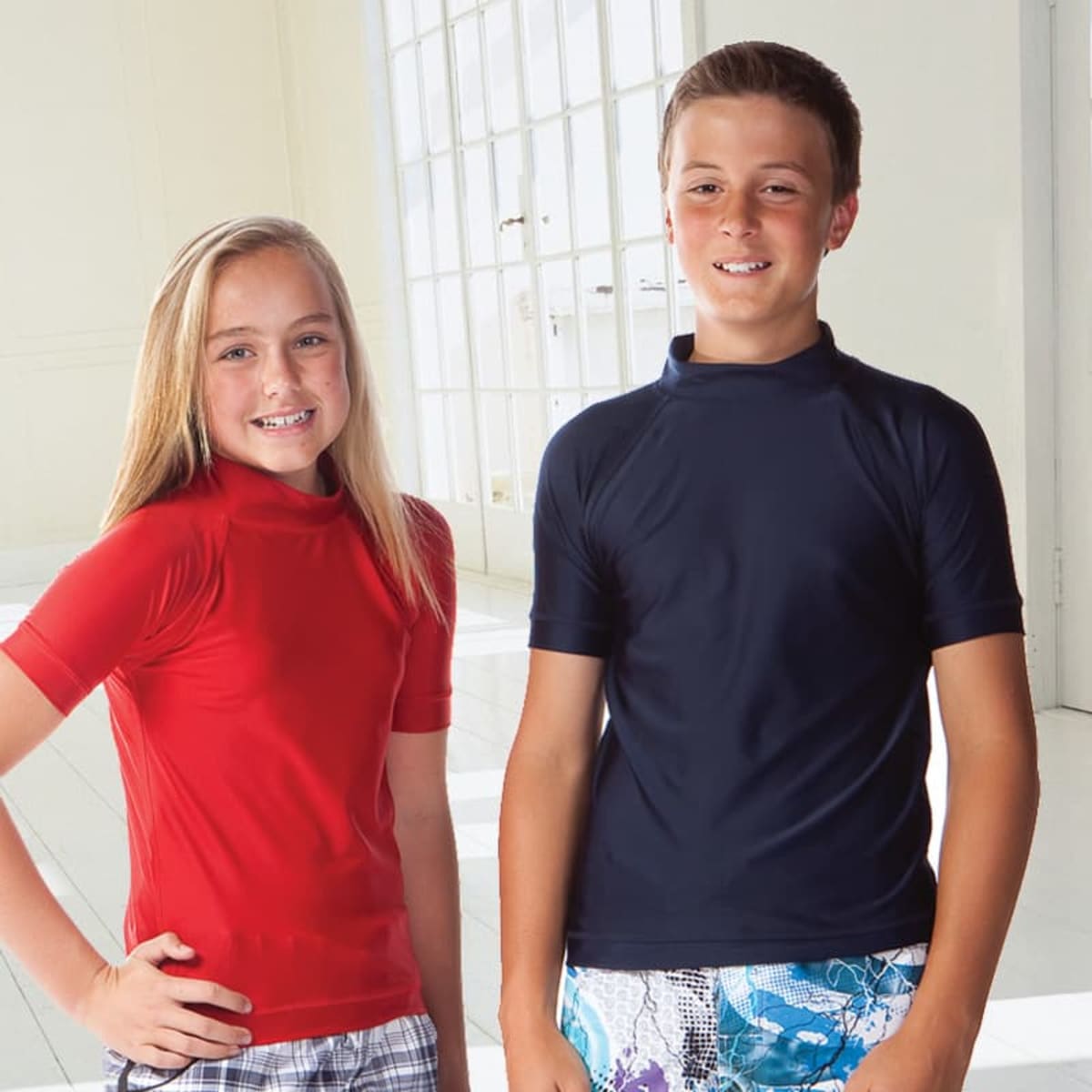 Two children are wearing rash vests; one in red and the other in navy blue, standing indoors with sunlight streaming in.