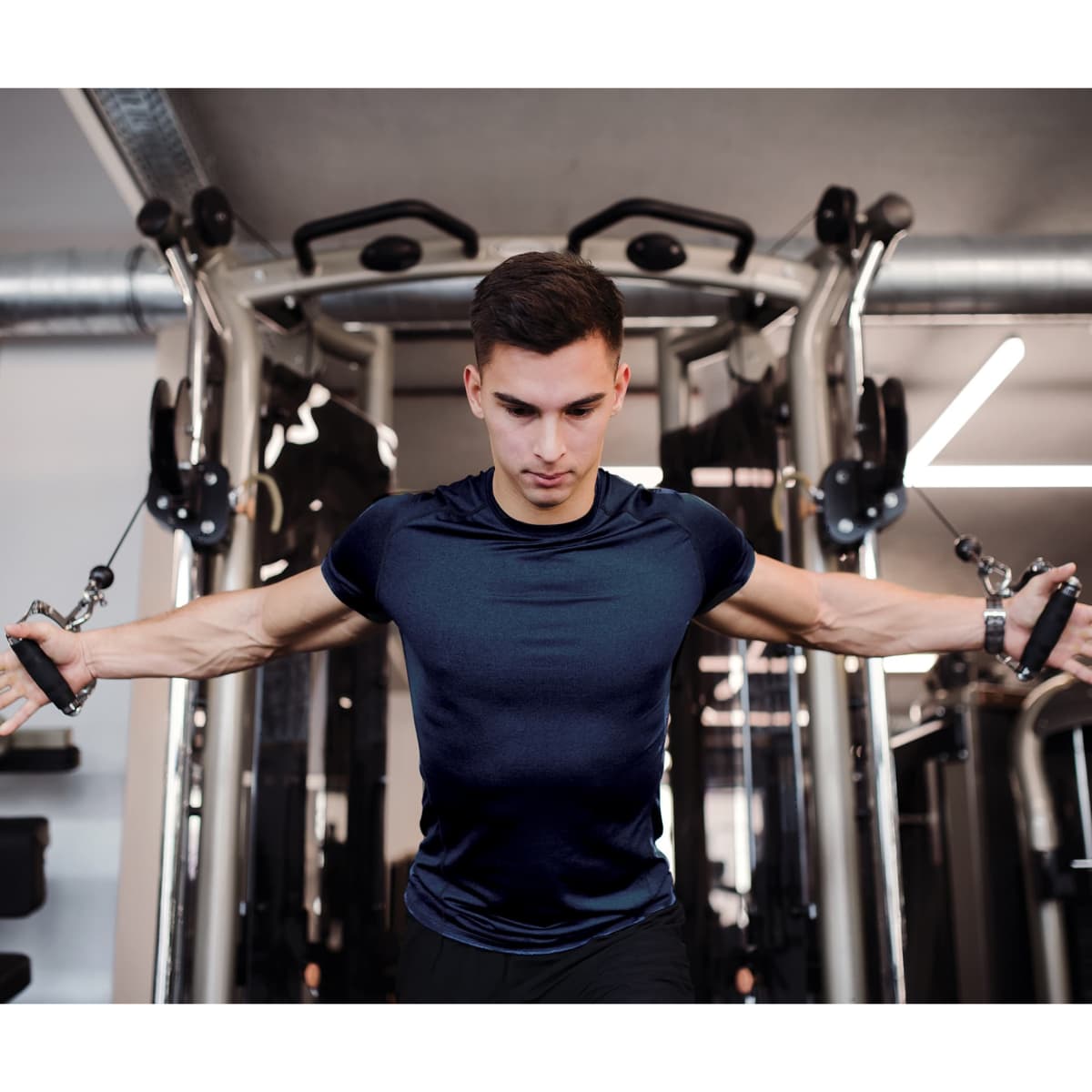 A fit man wearing a navy blue active tee exercises at a gym, using a cable machine. The setting features gym equipment.