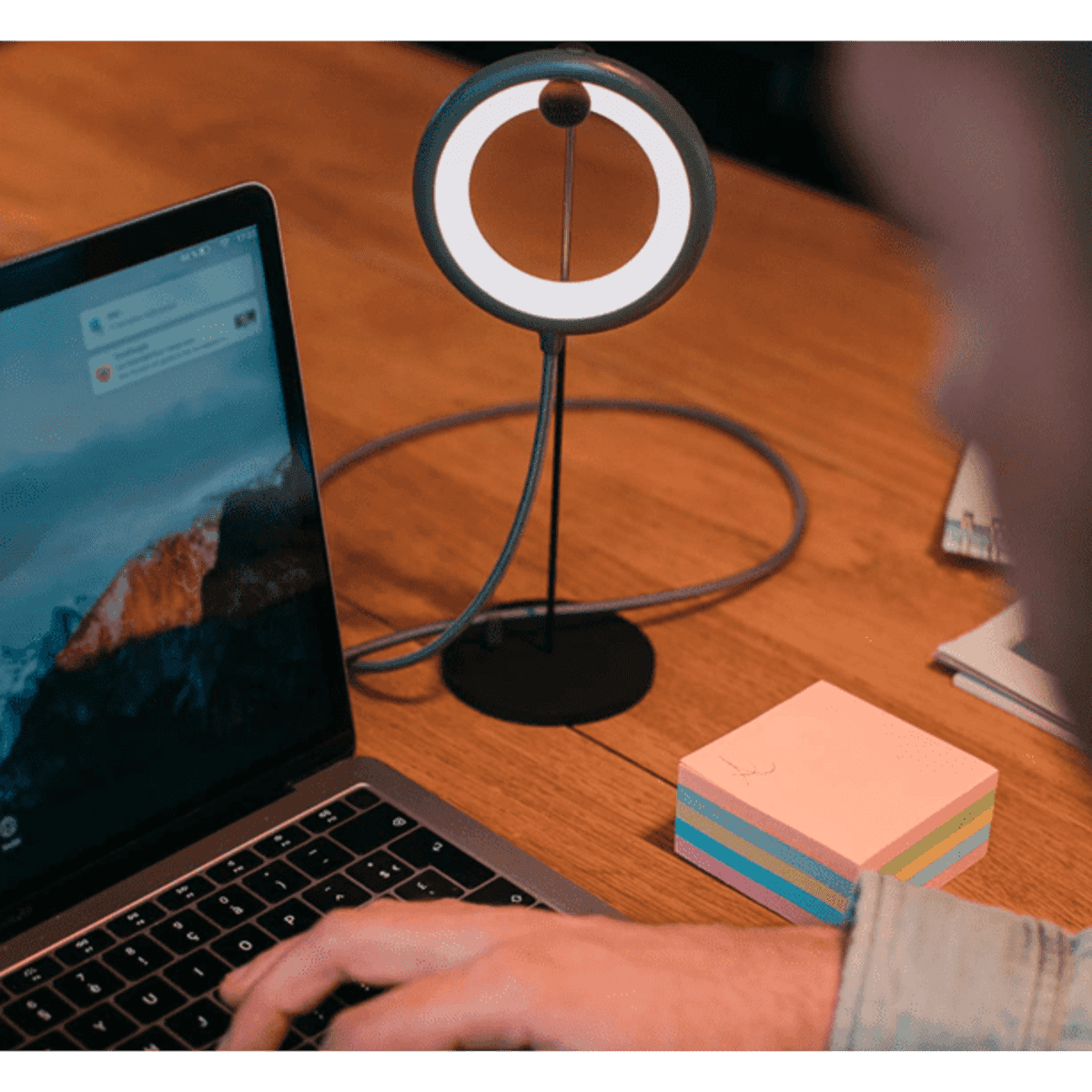 A sleek black lamp with a circular light stands beside a laptop on a wooden desk, with colourful sticky notes nearby.
