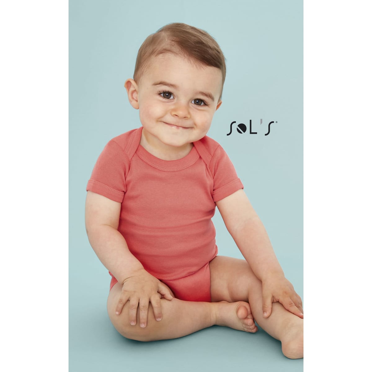 A baby bodysuit in coral pink, featuring short sleeves, displayed by a smiling child against a light blue background.