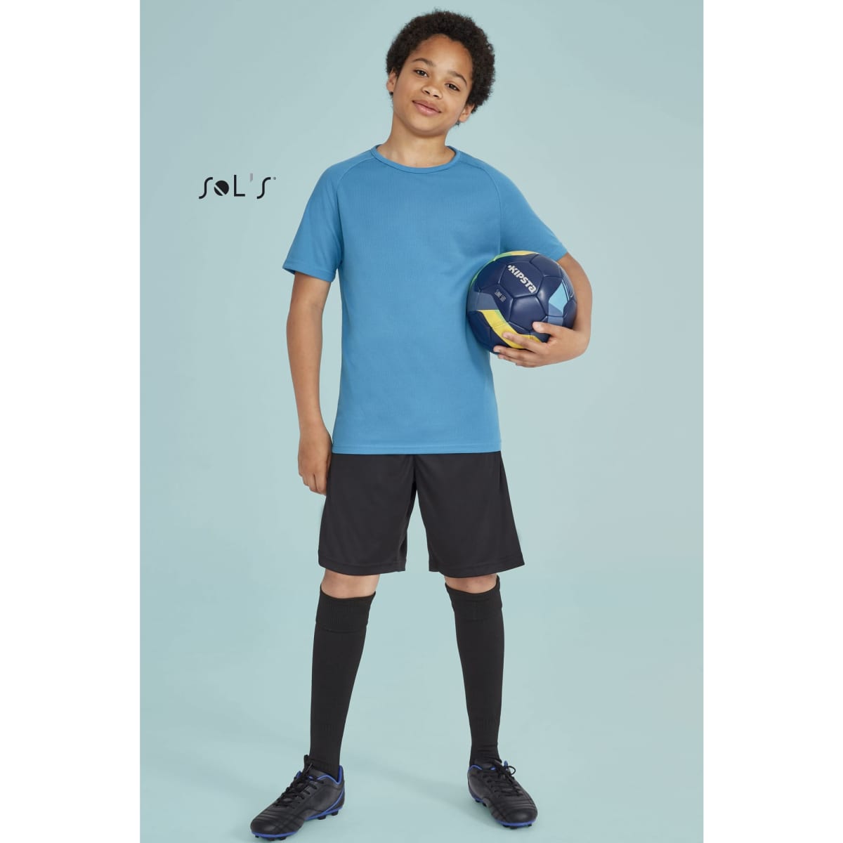 A boy stands holding a blue sport ball, wearing a blue breathable polyester T-shirt, black shorts, and black socks.