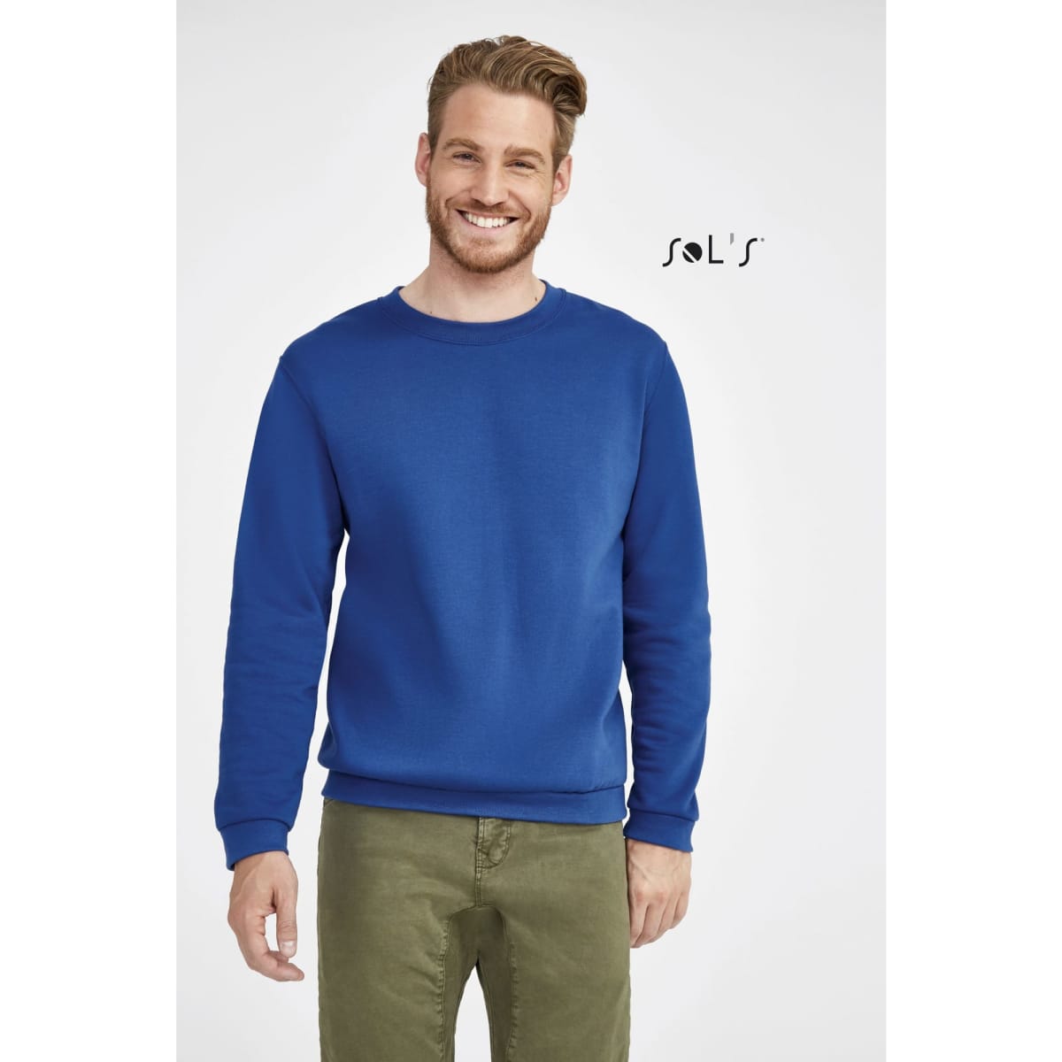 A man wearing a blue sweatshirt stands smiling, paired with olive green trousers. The sweatshirt features a logo.