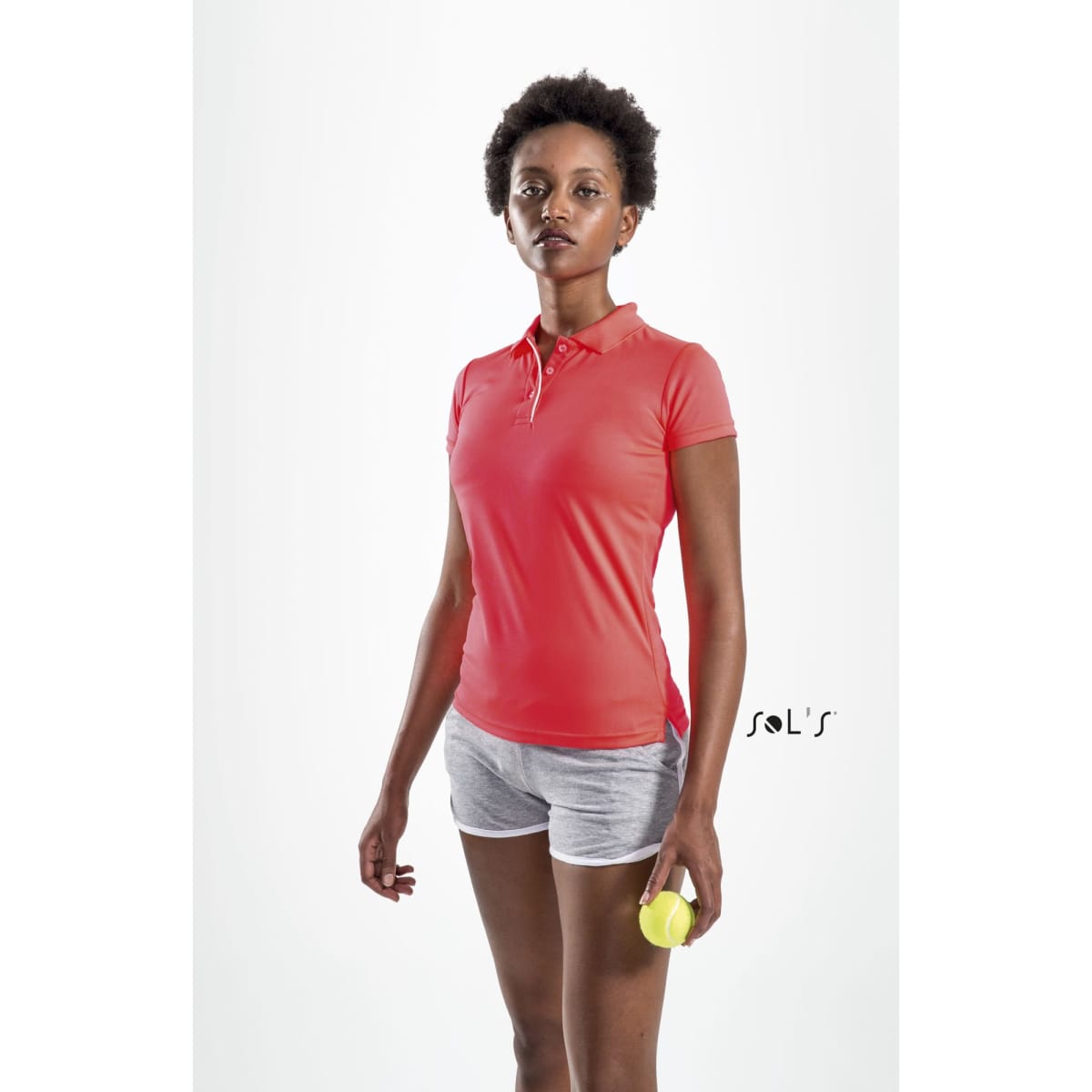 A woman wearing a pink polo shirt and grey shorts, holding a tennis ball, with a plain background.