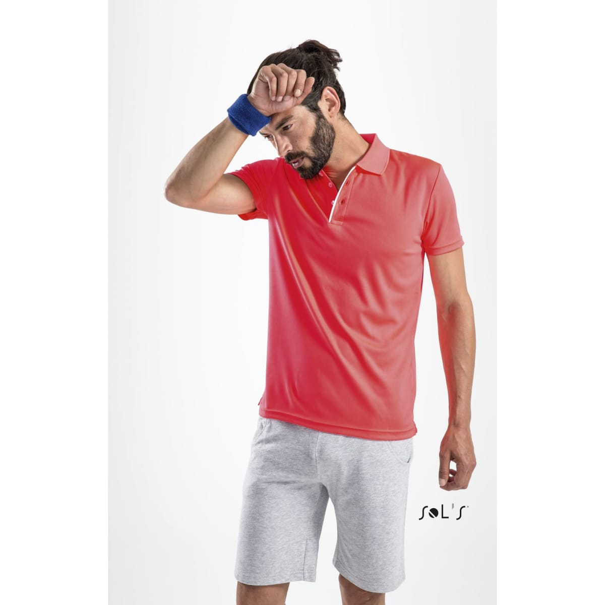 A man in a coral polo shirt and grey shorts wipes his forehead, featuring a small logo on the shirt.