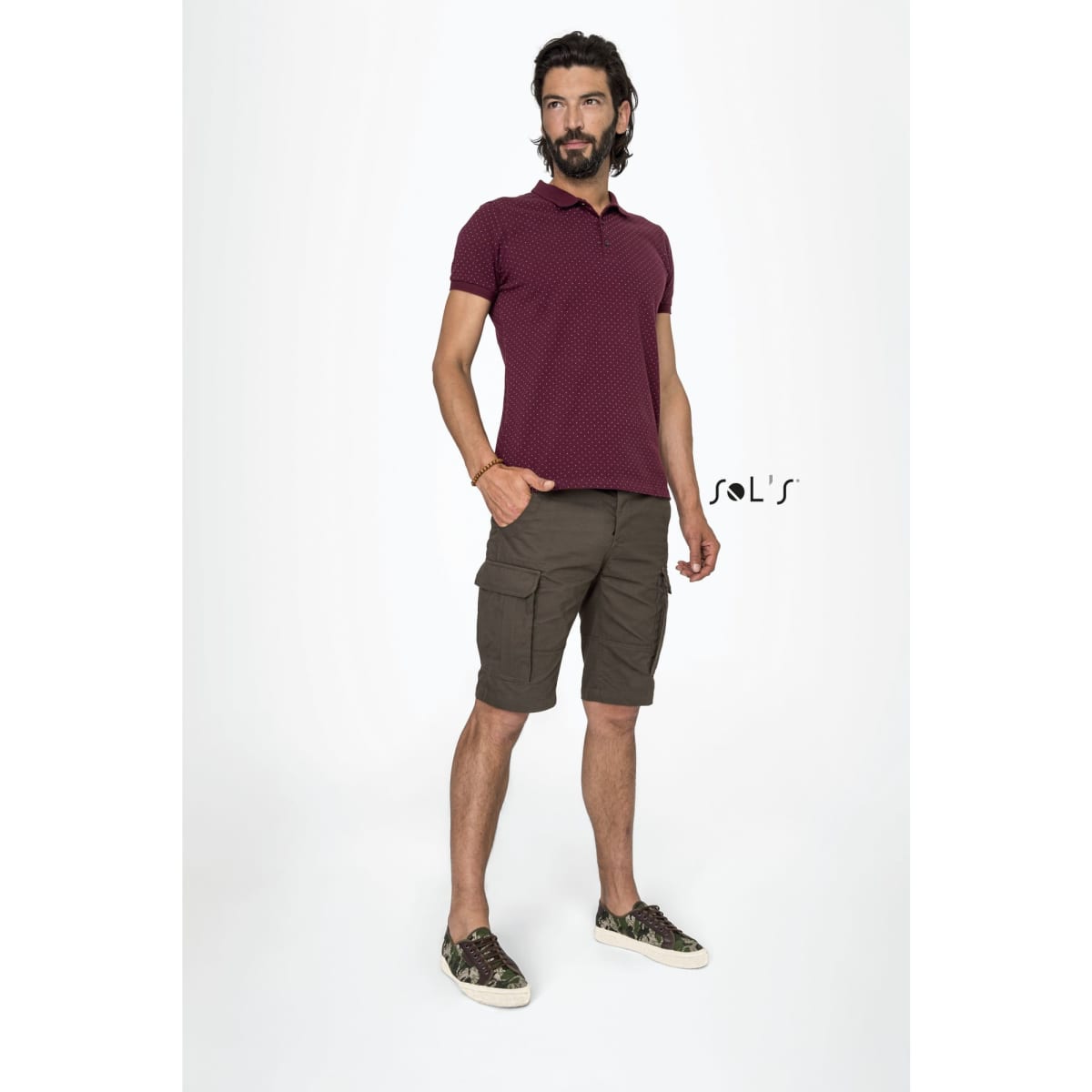 A man in a maroon polo shirt and brown Bermuda-style shorts, standing confidently with hands in pockets. The image includes a logo.