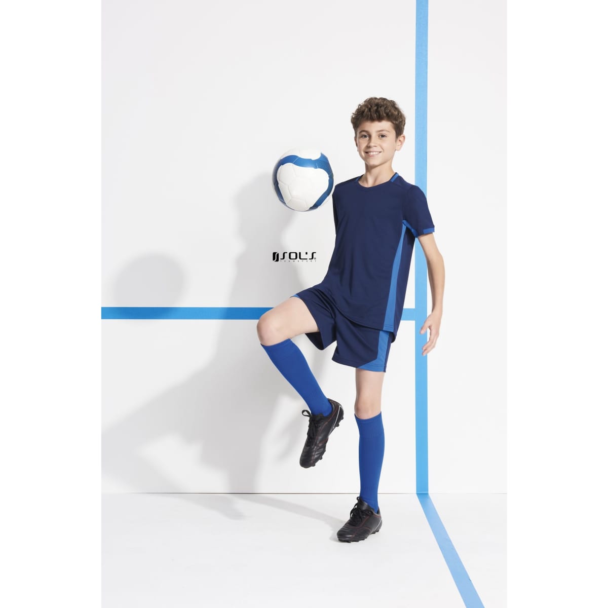 A child wearing a navy blue sports shirt and shorts, posing with a blue and white ball against a white backdrop.