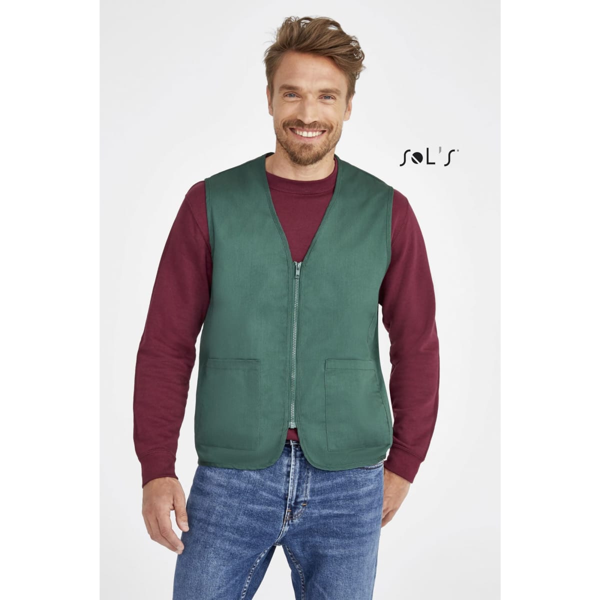 A green unisex zippered vest with two pockets, worn over a maroon long-sleeve shirt and denim jeans. It has a logo.