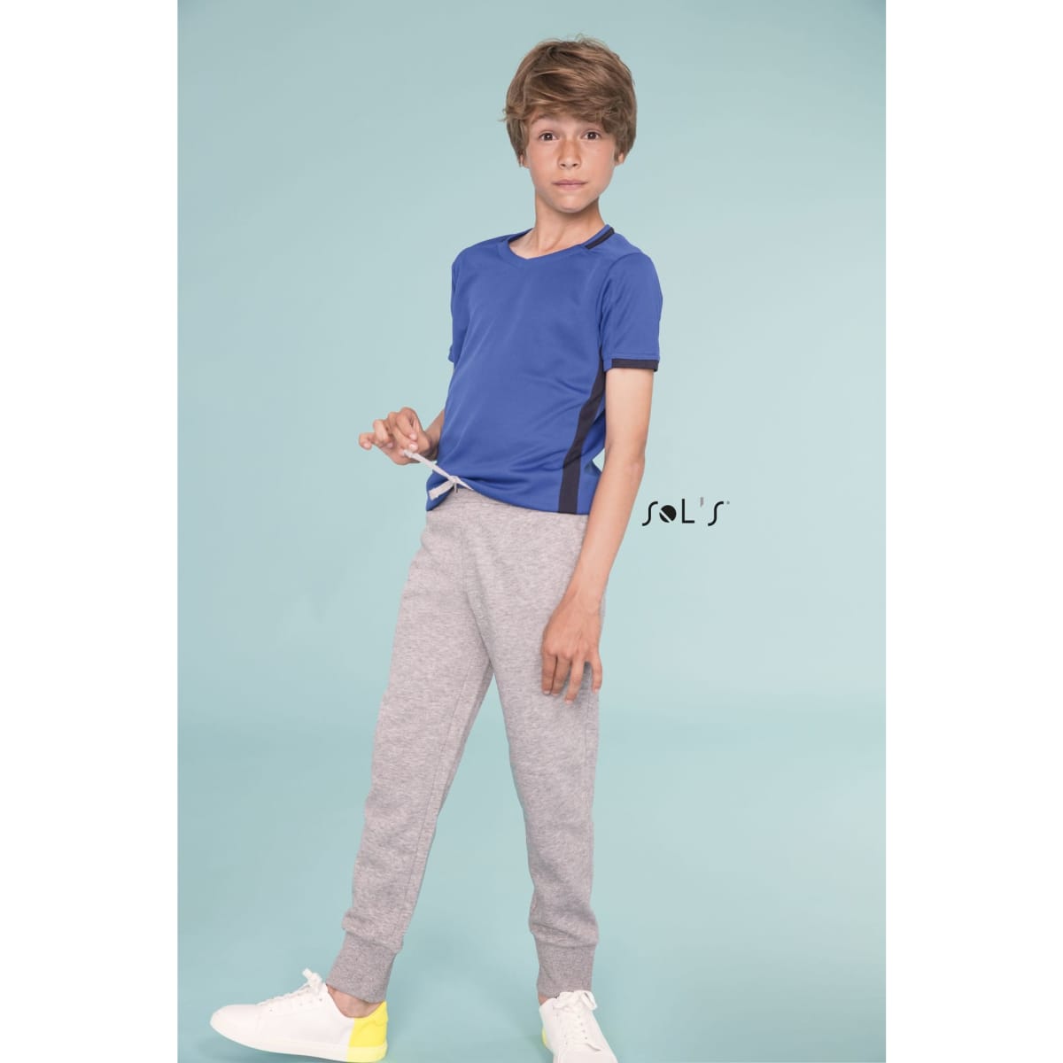 A boy in a blue t-shirt and grey slim fit track suit pants. The outfit features a logo.