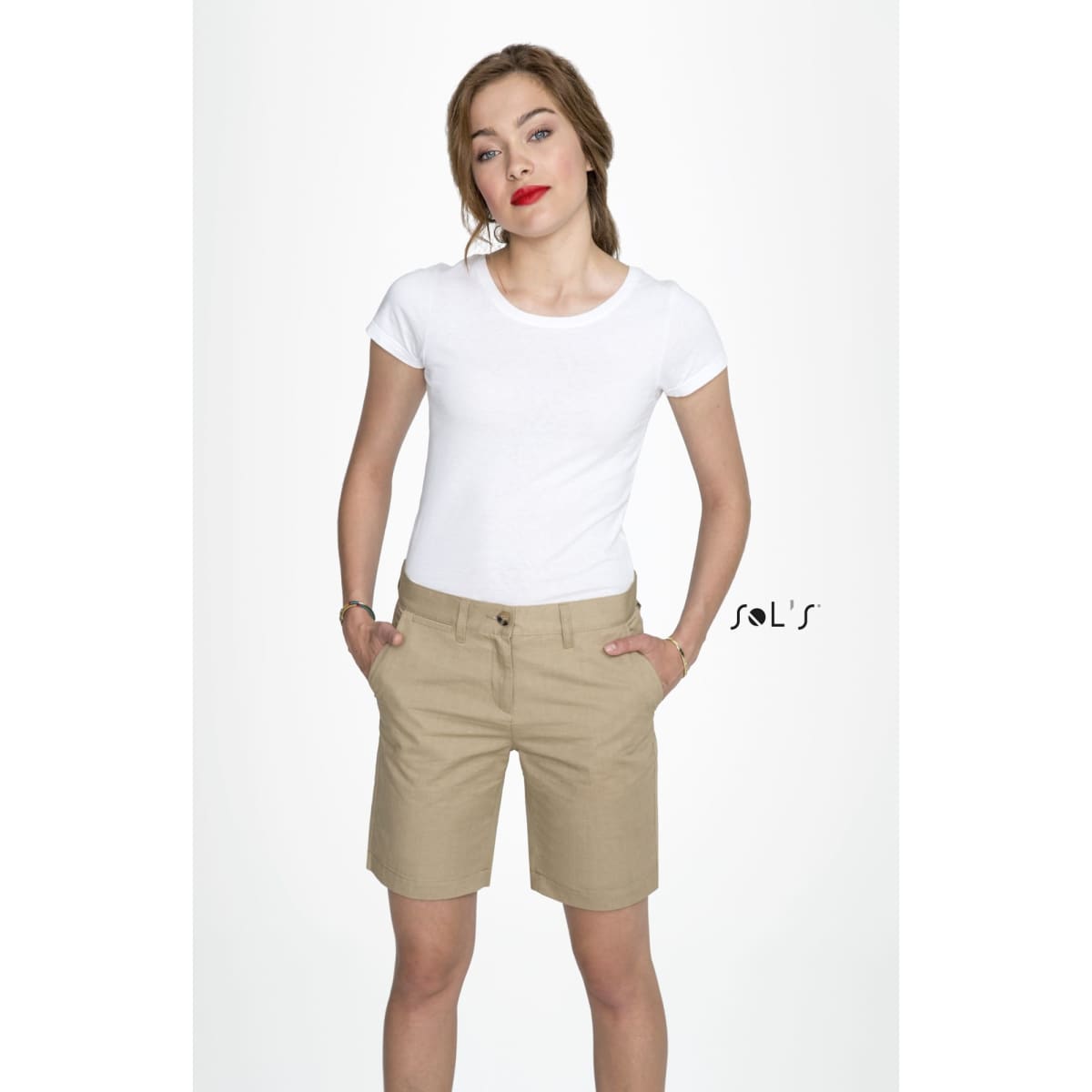 A woman models beige chino shorts with a white t-shirt, featuring pockets and a brand logo.