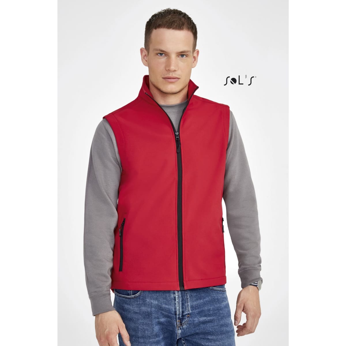 A red softshell vest with a high collar and zippered front, worn over a grey long-sleeve shirt. Features pockets and branding.