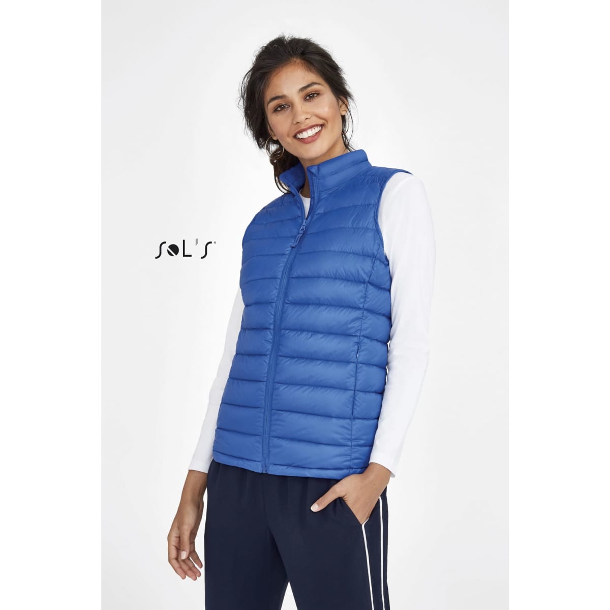 A woman wearing a blue puffer vest with a zip front, styled with a white top and dark pants, featuring a brand logo.