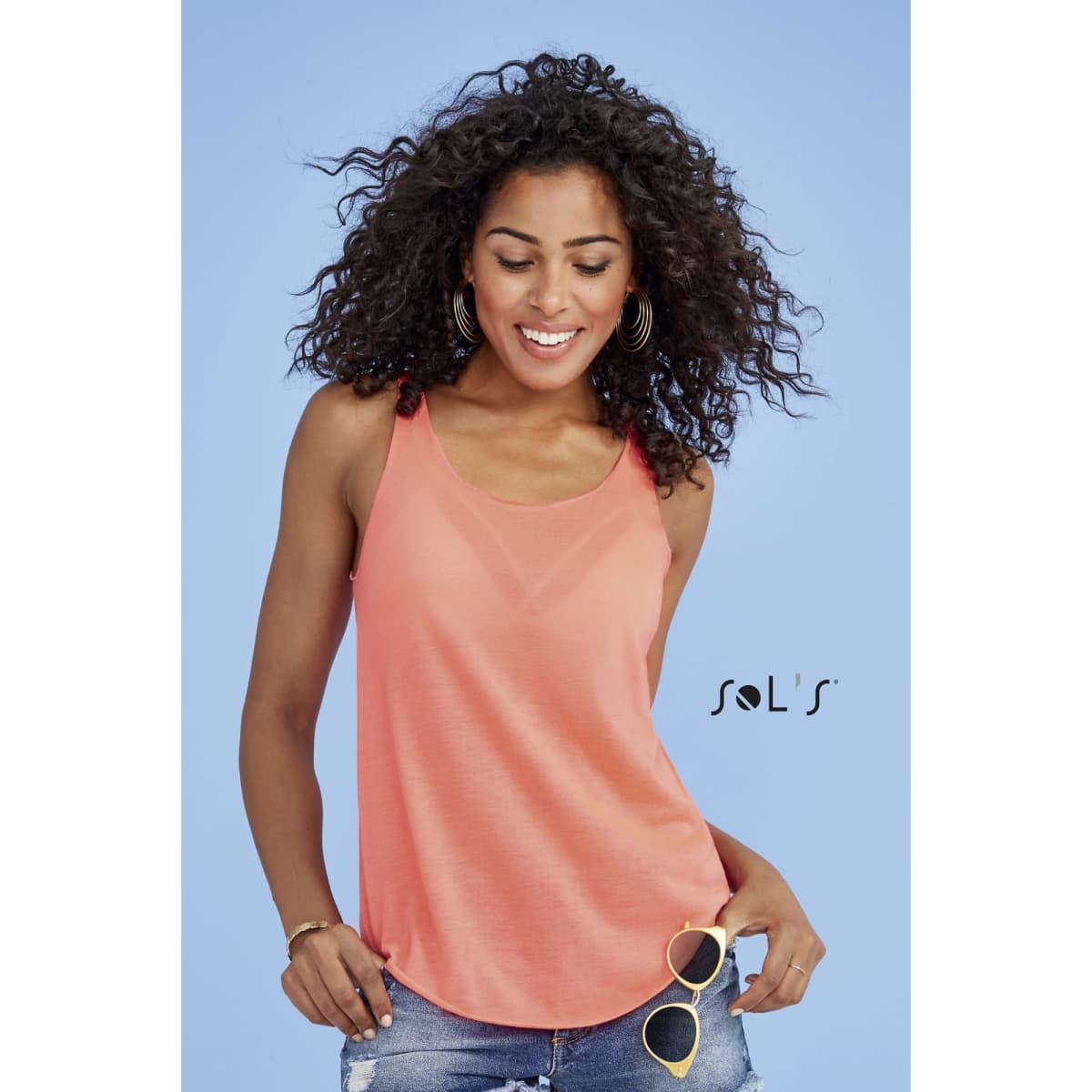 A woman wearing a coral tank top with a relaxed fit, paired with sunglasses and casual denim shorts. The product has a logo.