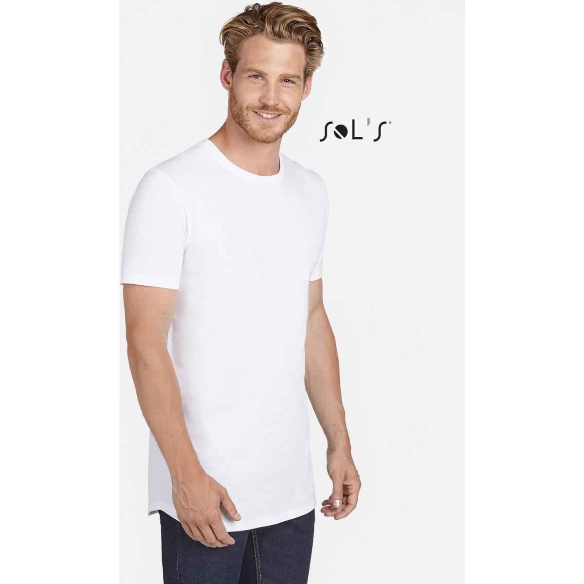 A man wearing a white long-length T-shirt, standing against a plain background with a logo in the corner.
