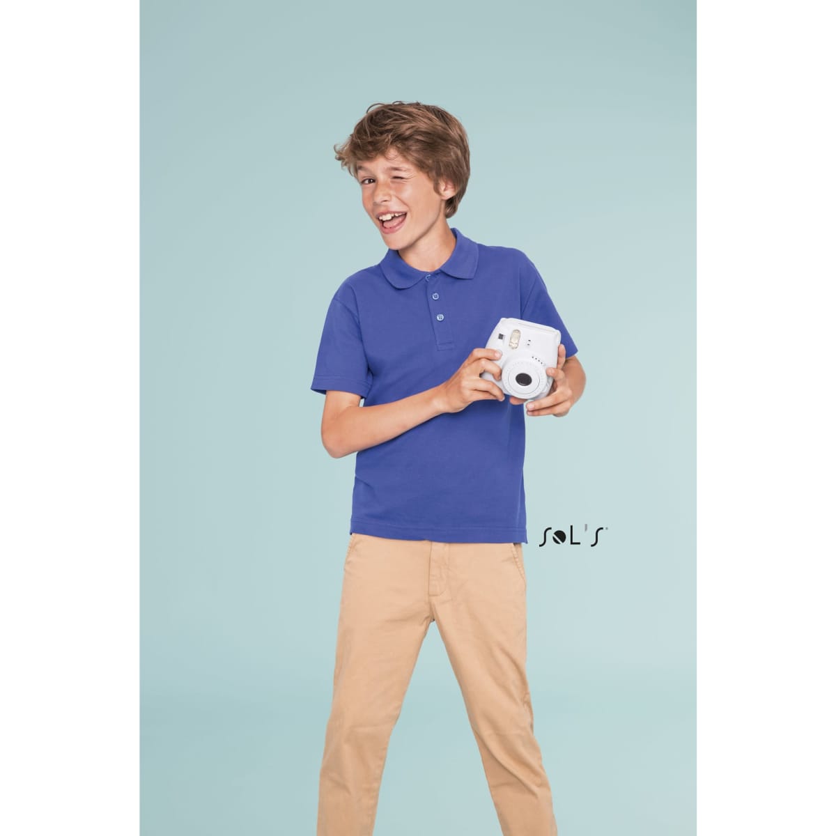 A boy in a blue polo shirt smiles while holding a camera, standing against a light background.