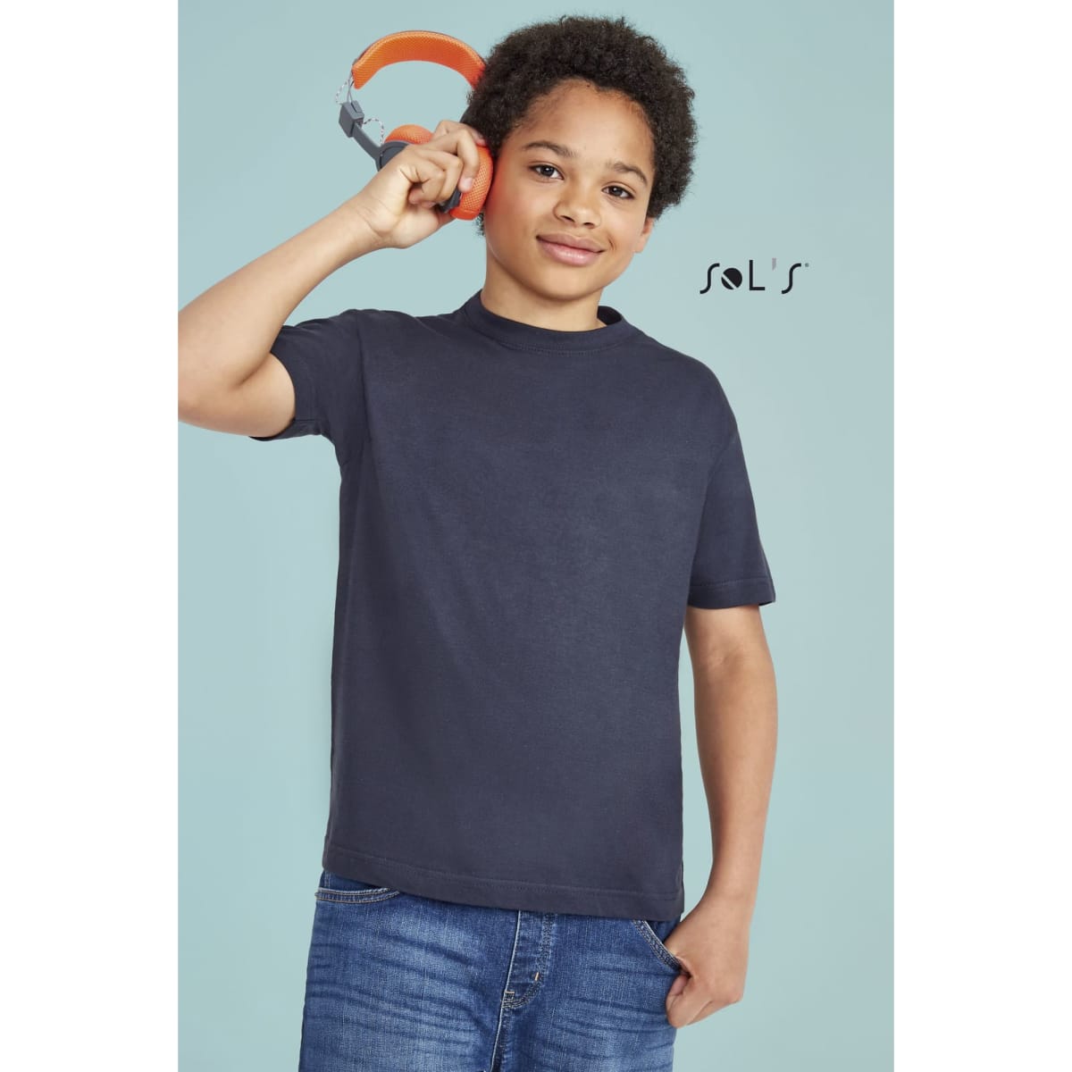 A navy T-shirt made from 100% semi-combed ring spun cotton, worn by a child holding headphones. Features branding.