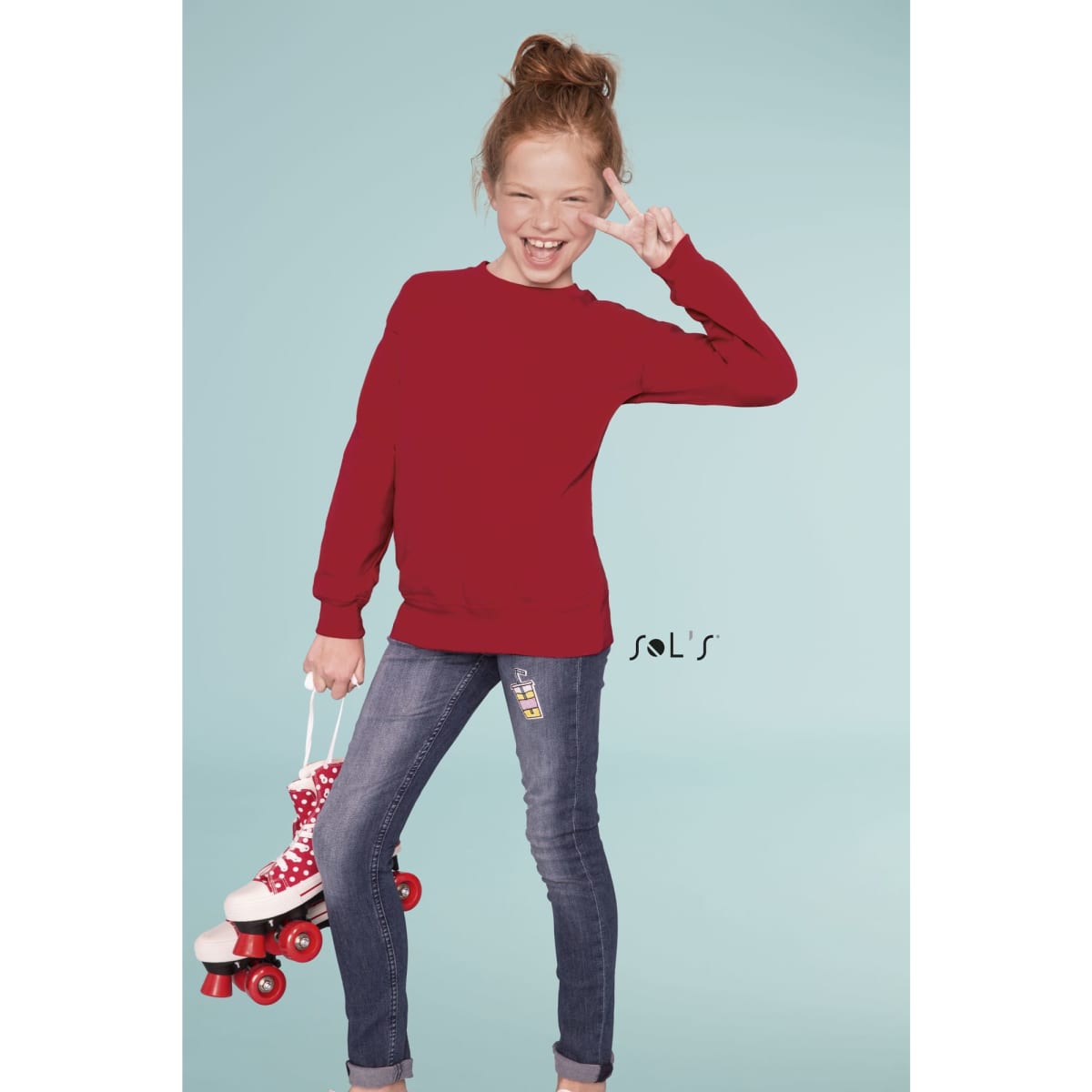 A girl in a red sweatshirt poses with roller skates while standing in a light blue background. The outfit features a logo.