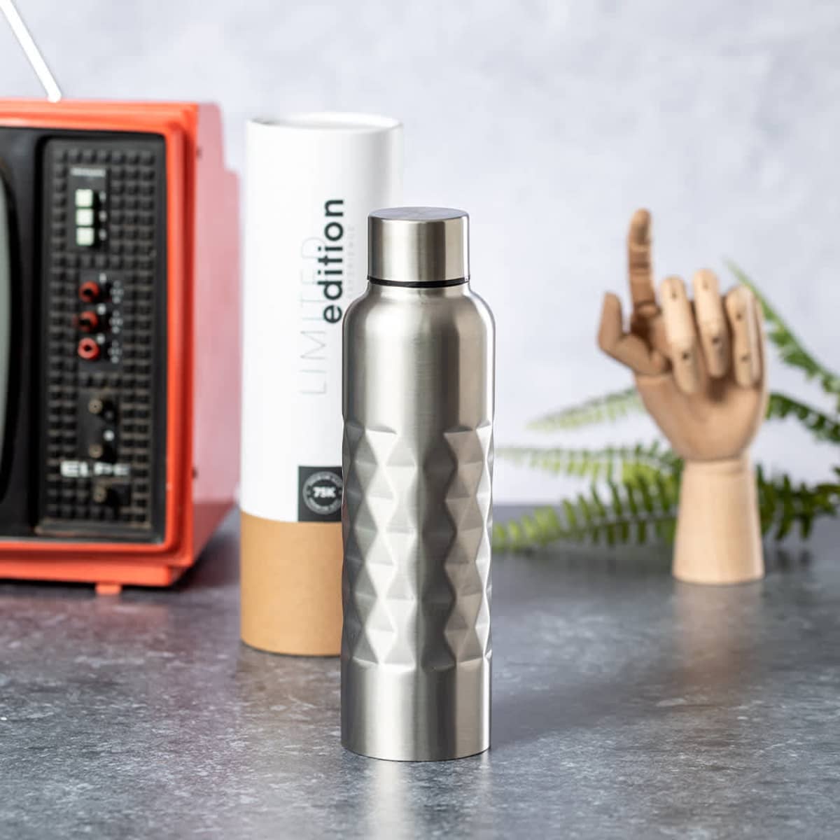 A drink bottle in stainless steel with a textured design, accompanied by a cylindrical container and a decorative hand model.