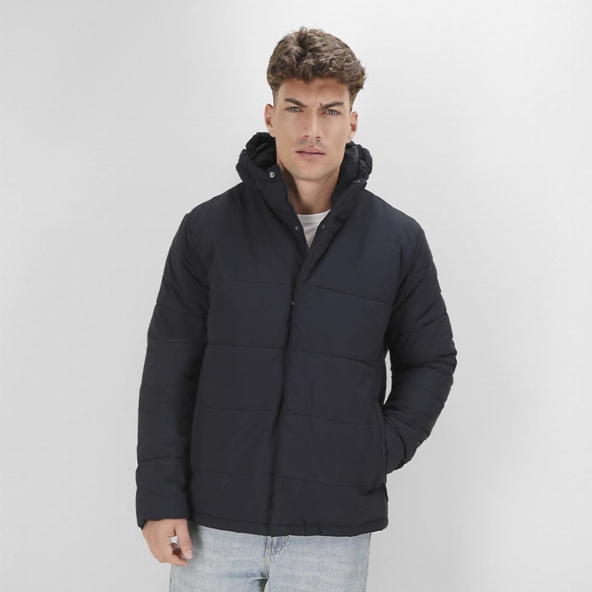 A man wearing a navy puffer jacket with a hood stands against a plain background.