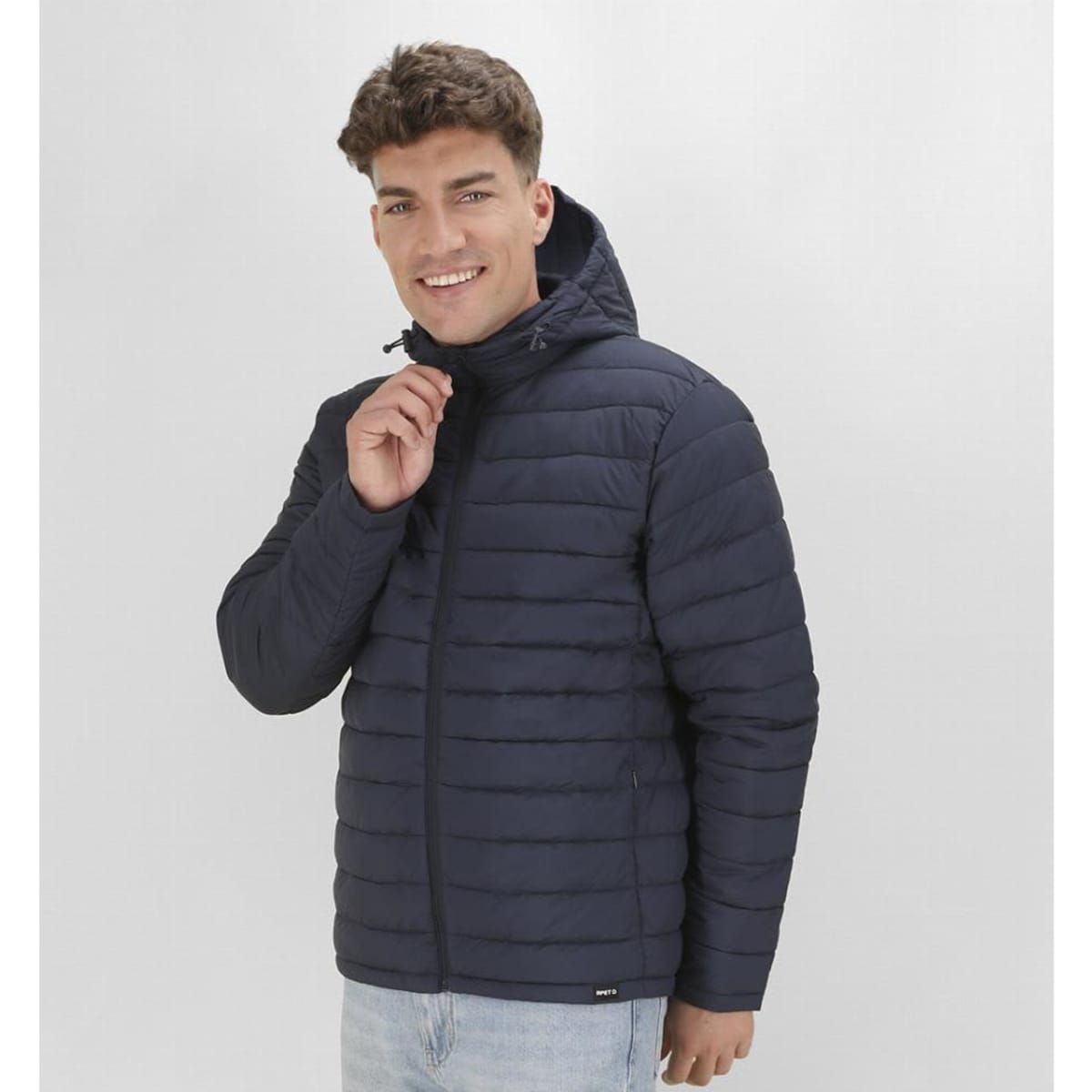 A man wearing a navy blue puffer jacket with a hood, smiling and adjusting the zipper.