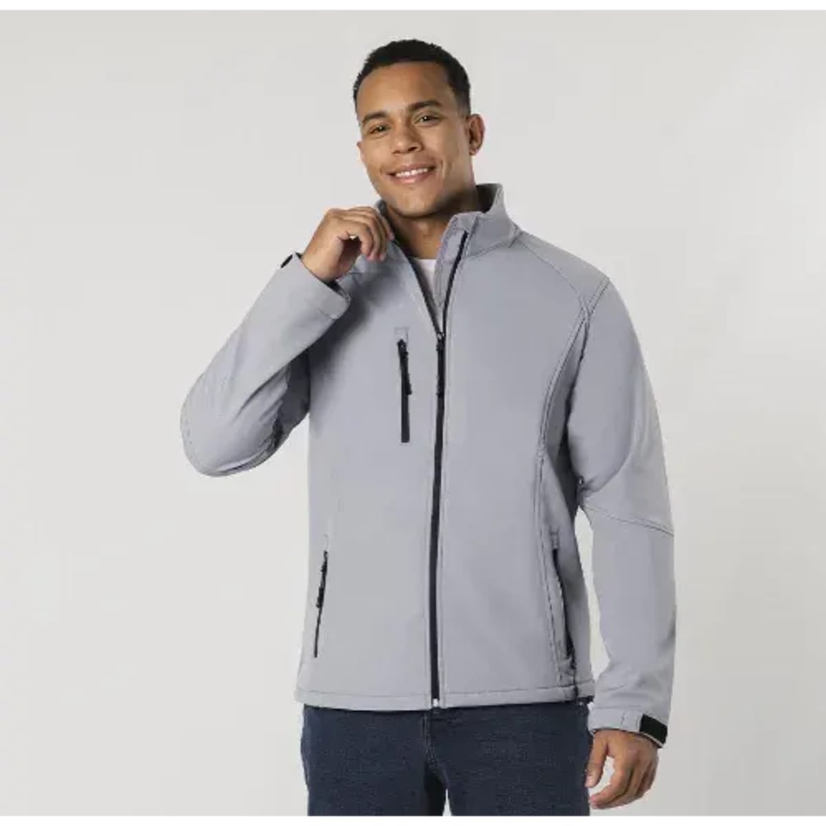 A man models a soft shell jacket in light grey, featuring a zippered front pocket and a collar.