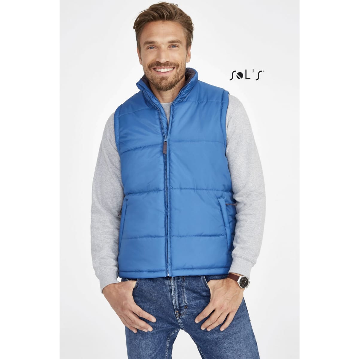 A man is wearing a blue puffer vest over a grey sweater, smiling while standing against a neutral background.