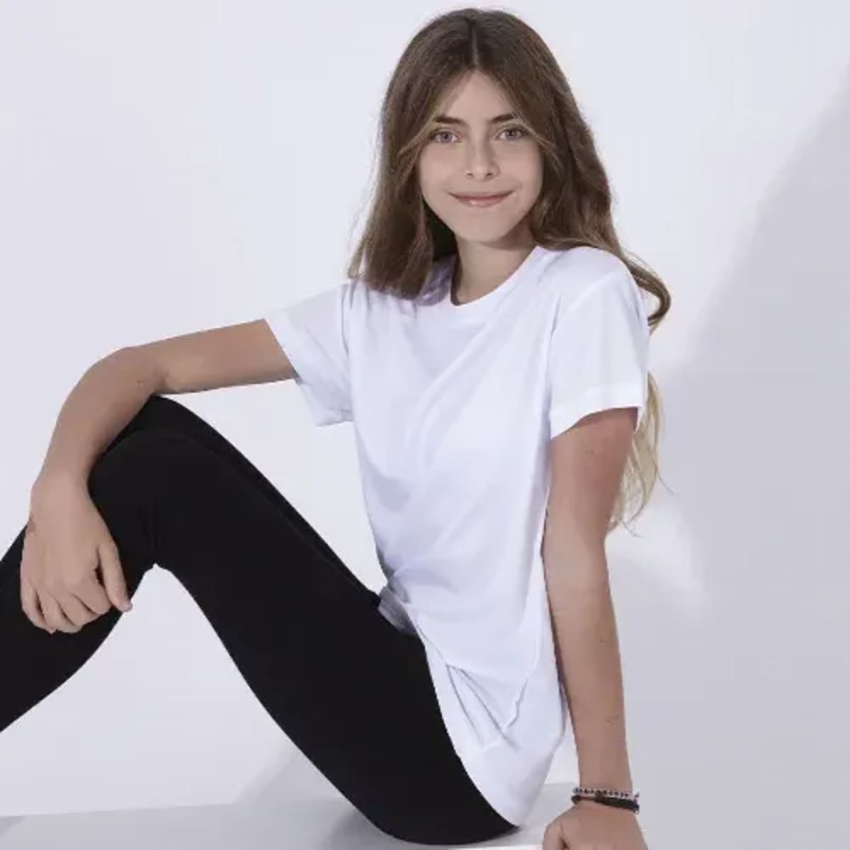 A white T-shirt is displayed, worn by a girl seated in black leggings, with long brown hair and a smile.