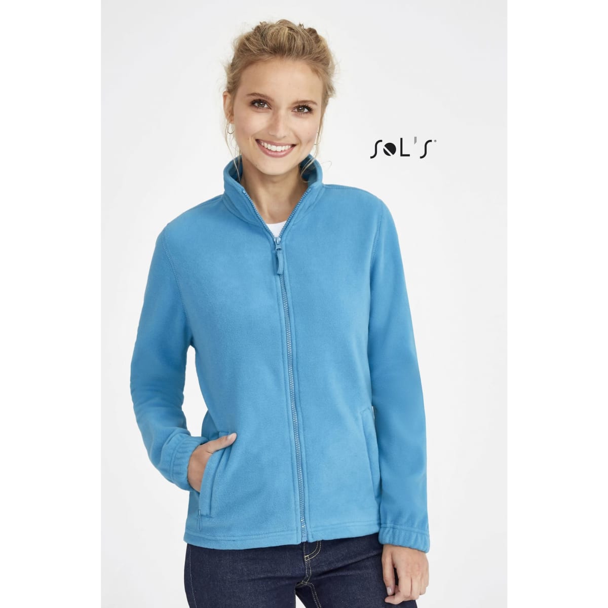 A woman models a zippered fleece top in light blue, featuring a collar and side pockets. The garment has a logo.