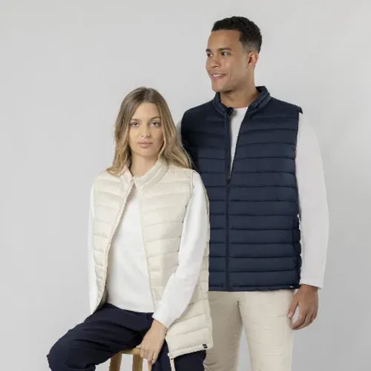 A cream puffer vest and a navy puffer vest worn by two models against a neutral background; both have logos.