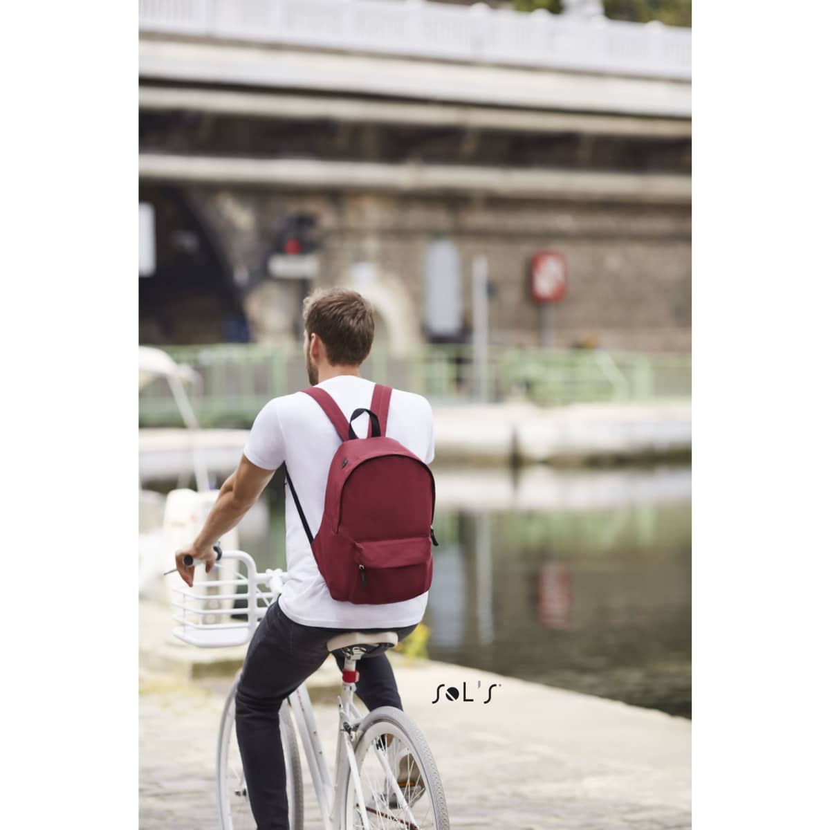 A burgundy rucksack with shoulder straps, placed on a cyclist's back, near a waterway. It features a logo.