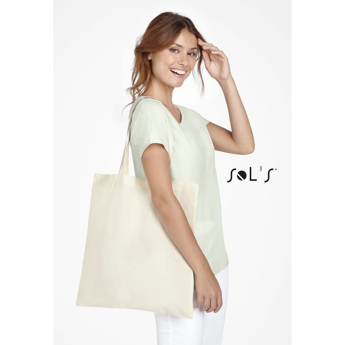 A person holds a natural beige tote bag, wearing a light green top and smiling, with a logo visible on the bag.