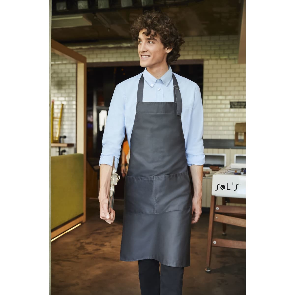A charcoal grey apron with pockets, made from polycotton, worn by a smiling man in a casual setting.