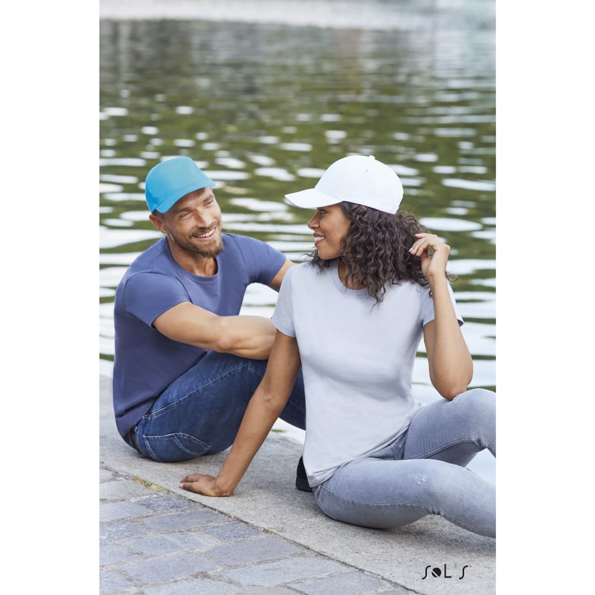 Two models by a water feature, one wearing a light blue cap and the other in a white cap, both in casual attire.