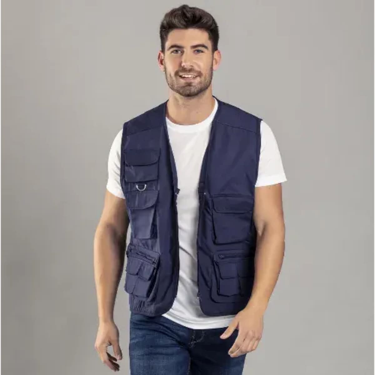 A man wearing a navy blue multi-pocket vest over a white t-shirt, standing against a grey background.