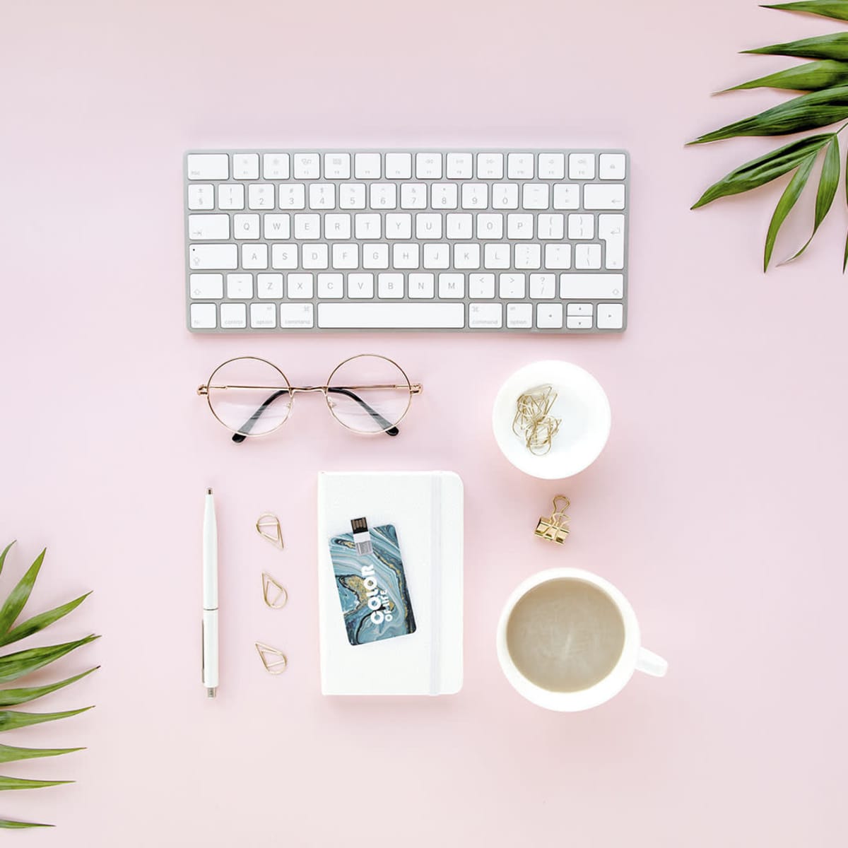 A flat lay of a stylish workspace featuring a white keyboard, glasses, a cup of coffee, and a USB memory stick.
