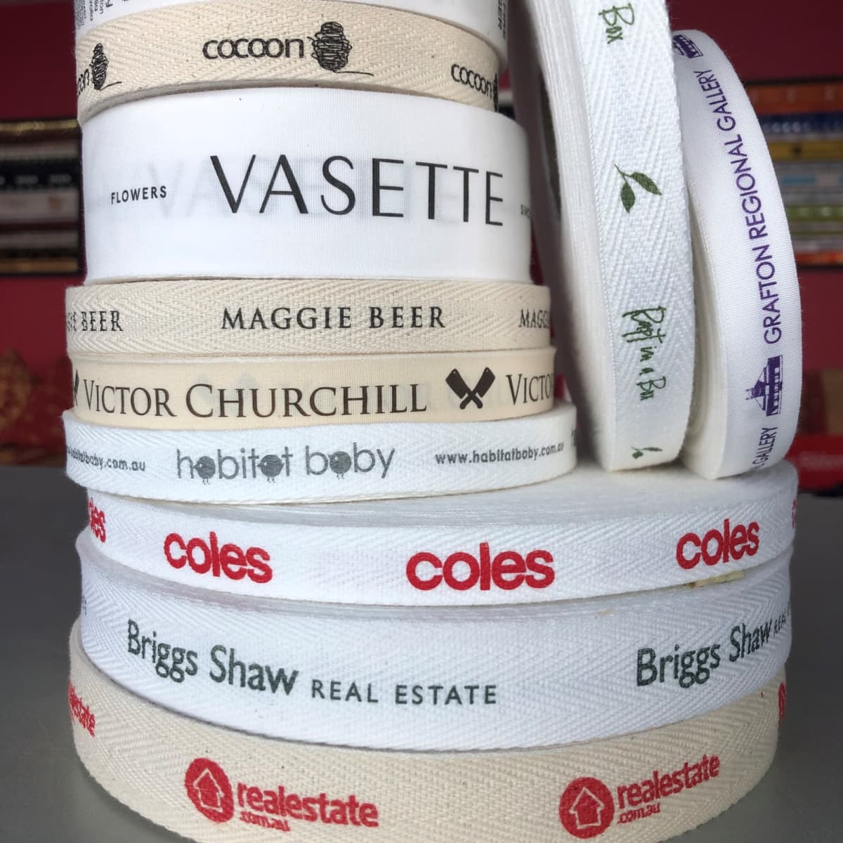 A stack of custom printed flat and herringbone cotton ribbon in cream and white featuring various logos.