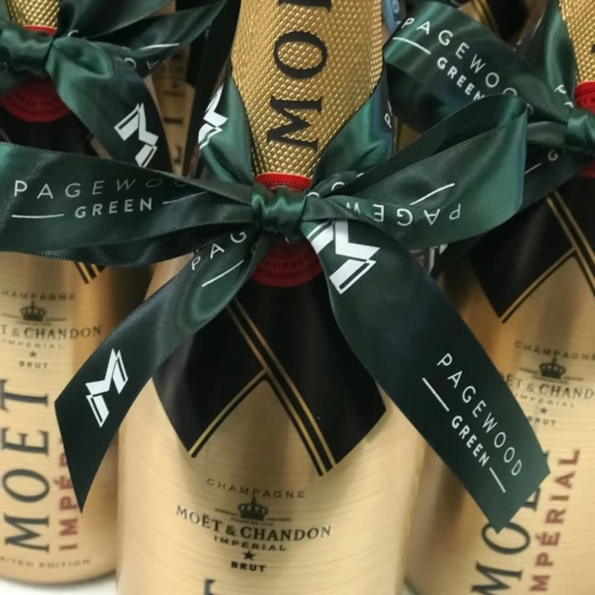 A gold champagne bottle wrapped with a dark green custom printed satin ribbon featuring a logo and text.