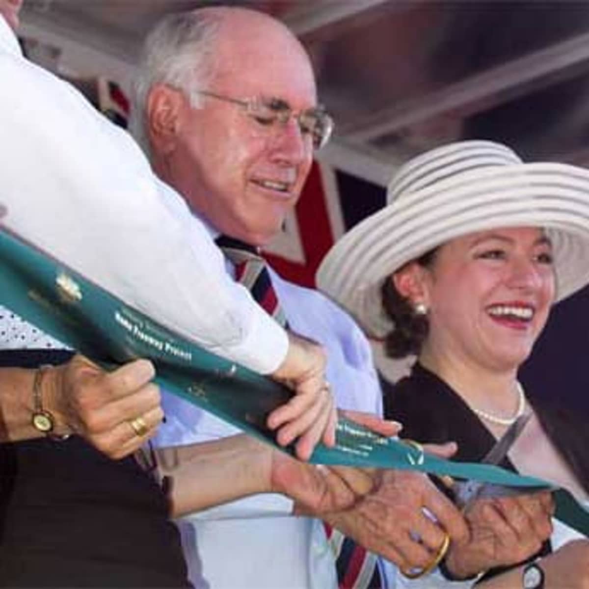 A green ribbon is being held by three individuals, with a focus on a man in a suit and a woman wearing a large hat.