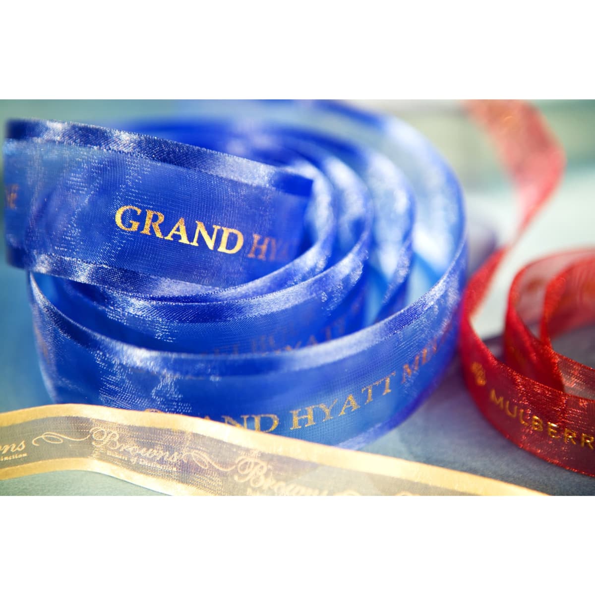 A roll of custom printed organza ribbon in blue, red, and gold, featuring different brands, with satin edges.