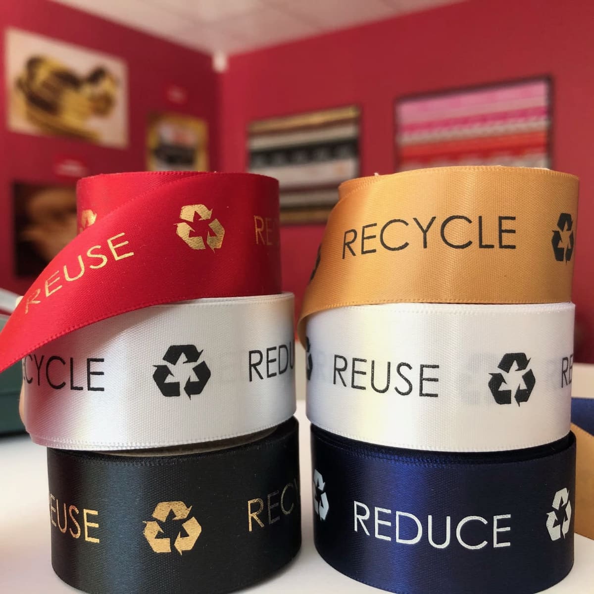 Custom printed recycled woven edge satin ribbons in red, gold, white, black, and navy, featuring eco-friendly logos.