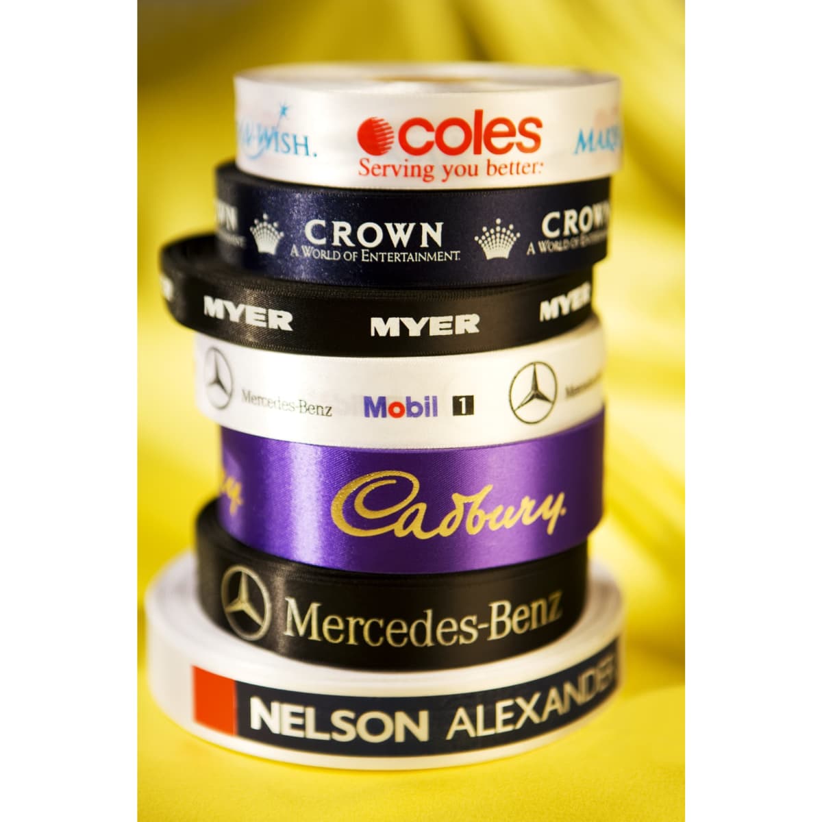A stack of various custom printed satin ribbons in multiple colours, featuring logos and branding.