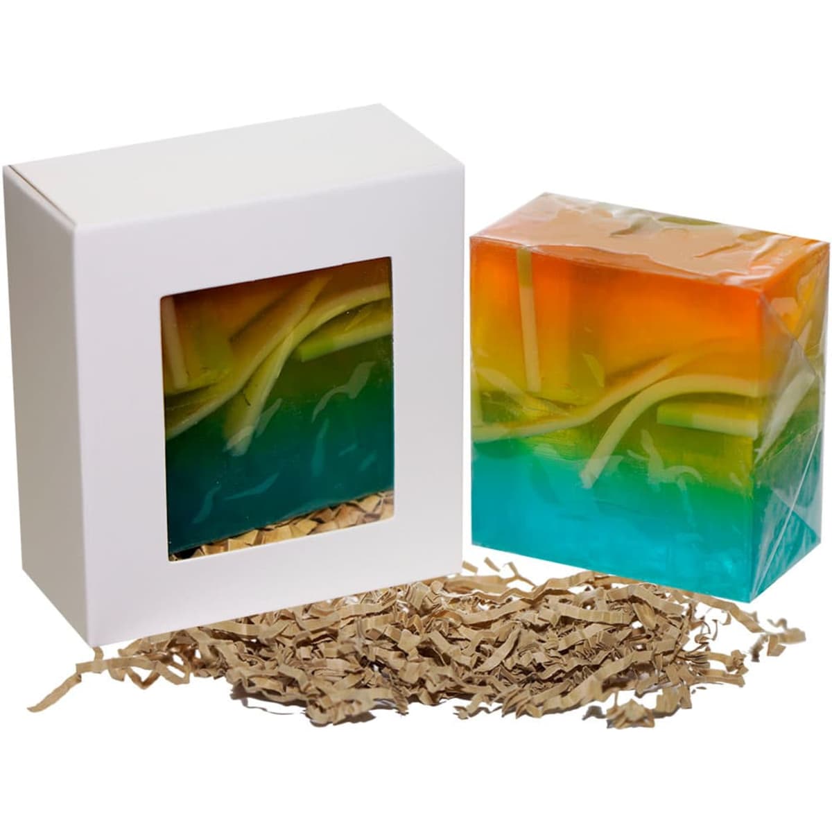 A colourful soap cube in orange and turquoise, presented in a white box with a clear window and shredded paper beneath.