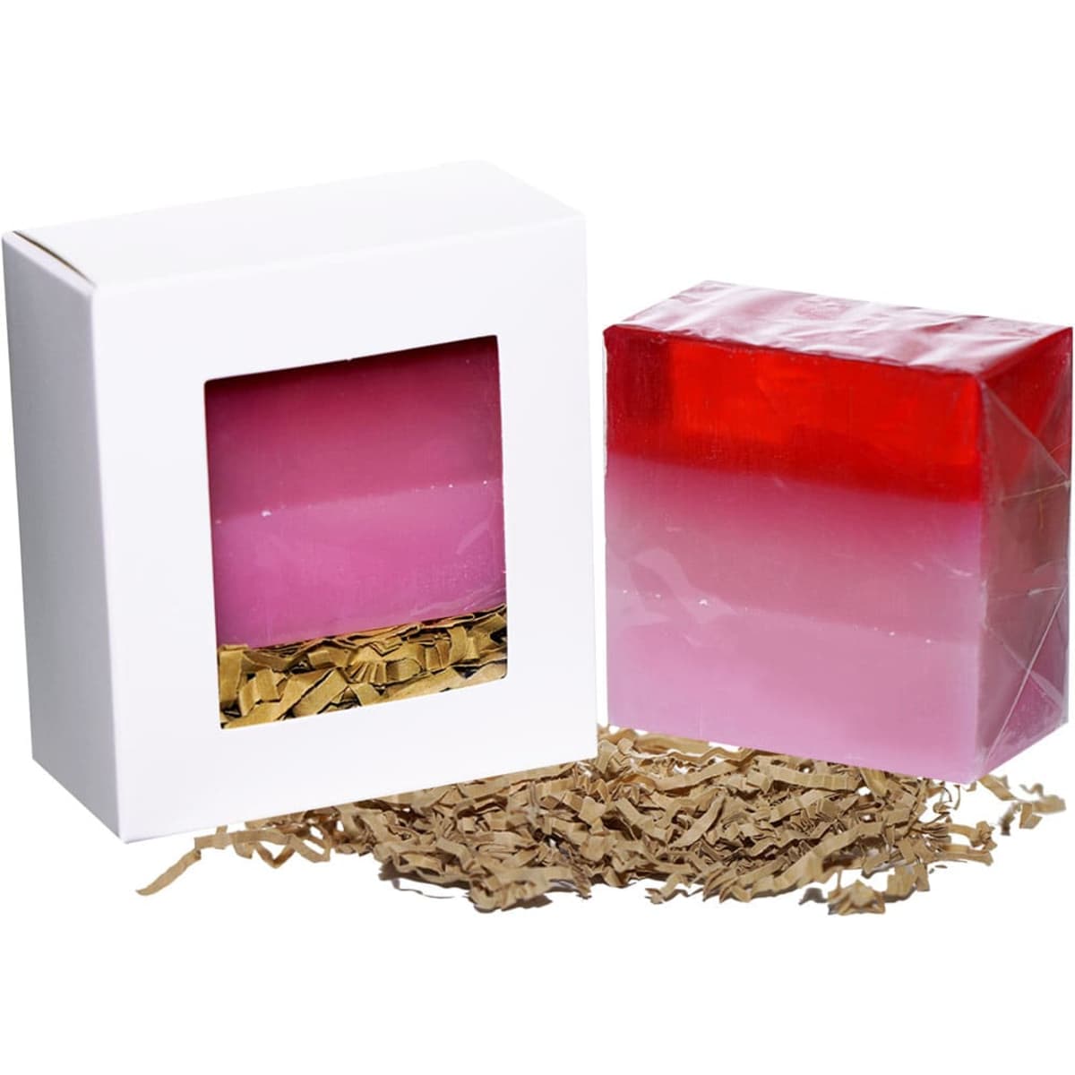 A pink and red layered soap bar beside a white box with a window, placed on decorative shredded paper.
