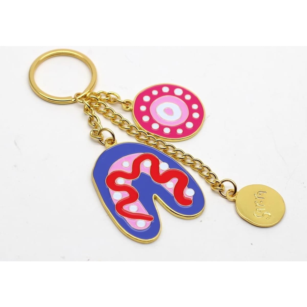 A colourful keyring featuring pink and blue shapes with red and white designs, attached to a gold chain and ring.
